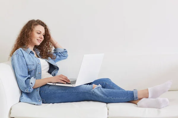 Horizontal shot of good looking curly haired young model sitting on white sofa, having laptop, watching videos, applying information, wearing jeans, white t shirt, socks and jacket. Tech concept. — Stock Photo, Image