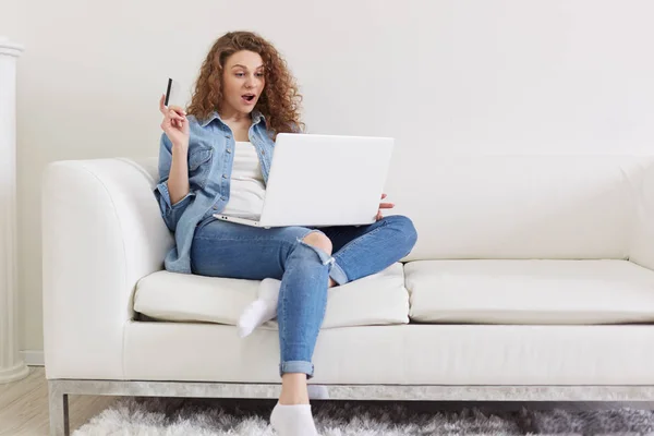 Indoor shot of shocked emotional lady with curly hair sitting in light living room, raising one hand, holding credit card, using her laptop, doing purchases on net, being fond of shopping online. — Stock Photo, Image