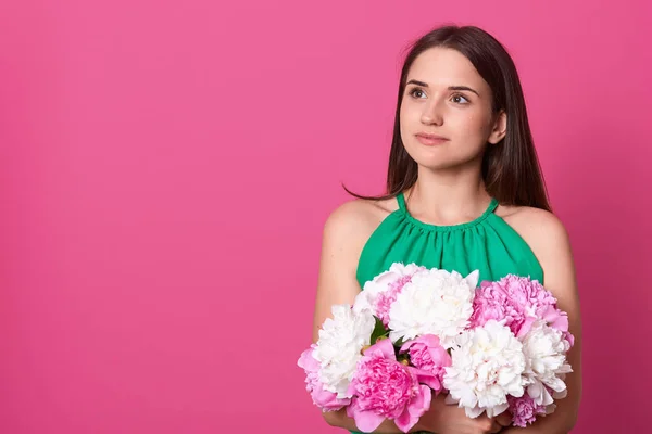 Imagen de la mujer joven soñadora reflexiva que tiene el pelo negro, mirando a un lado, teniendo mirada pensativa, usando vestido verde, sosteniendo flores coloridas, consiguiendo placer del regalo. Copyspace para publicidad . — Foto de Stock