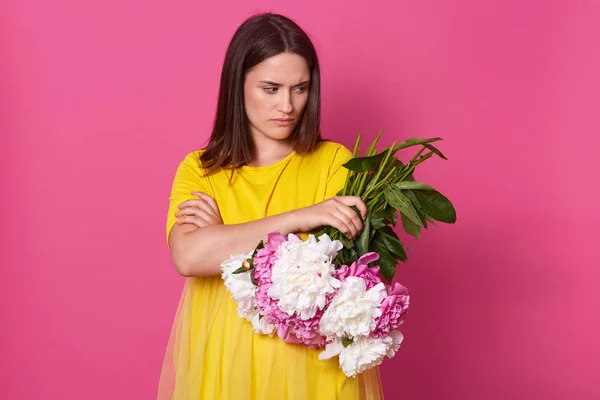 Разочарованная молодая женщина, стоящая со сложенными руками, держащая цветы в одной руке, глядя в сторону, с неприятным выражением лица, с короткими темными волосами, в ярко-желтом платье . — стоковое фото