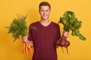 Happy cheerful young blond man holding healthy vegetables for salad. Raw foodist with carrots and beets posing isolated over yellow studio background. Raw dood diet and healthy eating concept. clipart