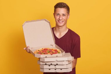 Yakışıklı genç teslim işçisi pizza kutuları yığını tutan, rahat tişört giyinmiş, kamera bakarak ve gülümseyen, lezzetli pepperoni ile açık kutu gösteren, sarı stüdyo arka planda izole poz