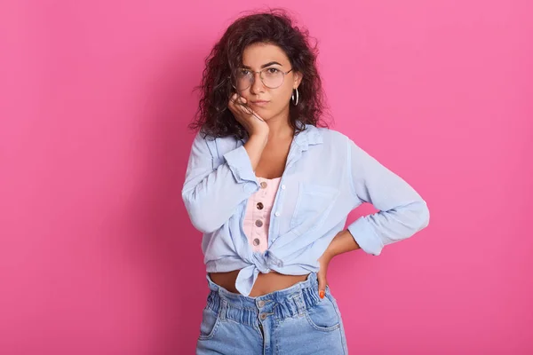 Mujer atractiva en gafas graduadas y atuendo de moda con expresión facial reflexiva, piensa en la pregunta, levanta las cejas, mantiene la mano bajo la barbilla, aislado en el fondo rosa. Concepto de personas . —  Fotos de Stock