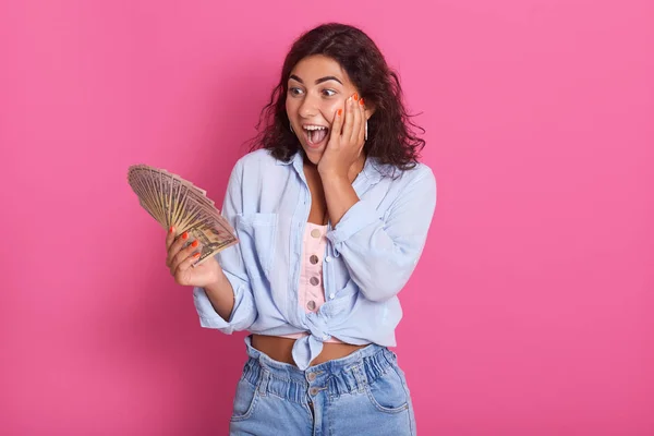 Porträtt av chockad ung brunett kvinna klädd i casual kläder, kvinnliga innehav fläkt av kontanter, har överraskat ansiktsuttryck, isolerade över rosa bakgrund i studion. Människor livsstil Concept. — Stockfoto