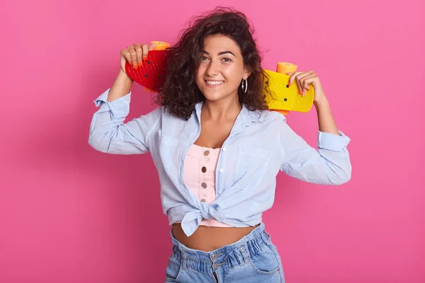 Närbild av brunett charmig kvinna med anbud leende, hålla skateboard på axlarna, klädd i t-shirt, skjorta och jeans, åtnjuter hobby, isolerade över rosa bakgrund, redo att åka skridskor i parken. — Stockfoto