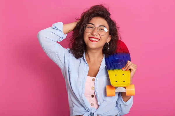 Primer plano retrato de la joven alegre en ropa casual de pie, mirando directamente a la cámara, la celebración de monopatín multicolor aislado sobre fondo rosado en el estudio. Concepto de vida . — Foto de Stock