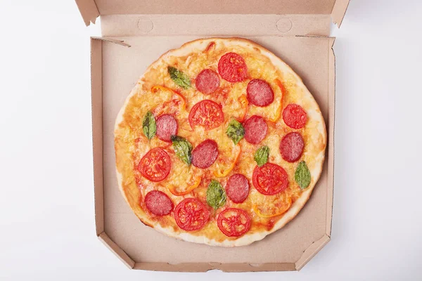 Pemandangan atas pizza panas lezat dalam kotak dengan ham dan tomat diiris dan disajikan di atas meja putih, makan malam lezat untuk pecinta makanan cepat saji di permukaan cahaya, kotak dengan pizza terisolasi di atas latar belakang putih . — Stok Foto