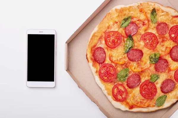 Pizza dalam kotak dan smartphone dengan layar kosong di atas meja putih, top view pepperoni lezat diisolasi di atas latar belakang cahaya. Ponsel modern dekat kotak dengan pizza. Salin ruang untuk advertismen . — Stok Foto