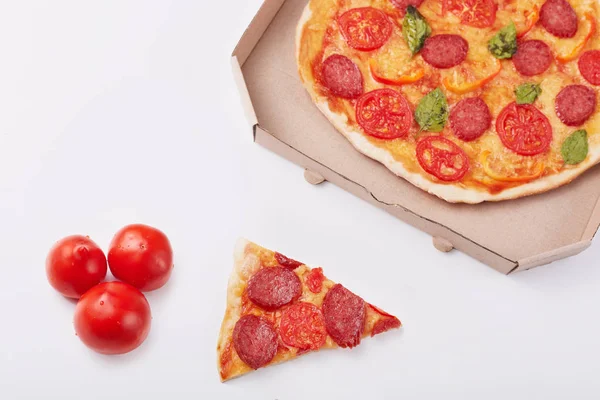 Photo of pepperoni pizza with mozzarella cheese, salami, pepper, spices and fresh spinach, cardboard box, piece of pizza and three tomatoes isolated on white background. Fast food eating concept. — Stock Photo, Image