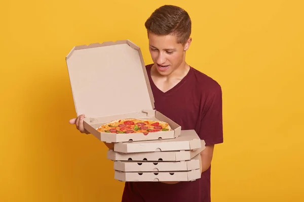 Foto hirizontal de un hombre guapo con una camiseta casual de color burdeos, sosteniendo una pila de cajas de pizza en las manos, mirando a la caja abierta, posando aislado sobre el fondo amarillo del estudio. Concepto de comida rápida . — Foto de Stock