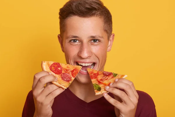 Foto de un tipo vestido con camiseta borgoña sosteniendo dos piezas de pizza y come comida rápida, disfruta de un delicioso plato, macho posando sobre el fondo amarillo del estudio, mirando a la cámara con expresión feliz . — Foto de Stock