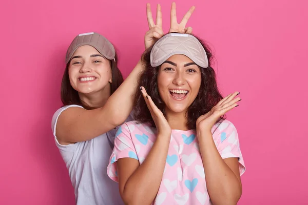 Χαρούμενες χαρούμενες κοπέλες με πιτζάμες και μάσκες ύπνου, έχουν και τα δύο χέρια πάνω από τη μάσκα και διασκεδάζουν δείχνοντας τη γλώσσα τους. Στούντιο κοντινό πορτραίτο σε απομονωμένο λευκό φόντο. — Φωτογραφία Αρχείου