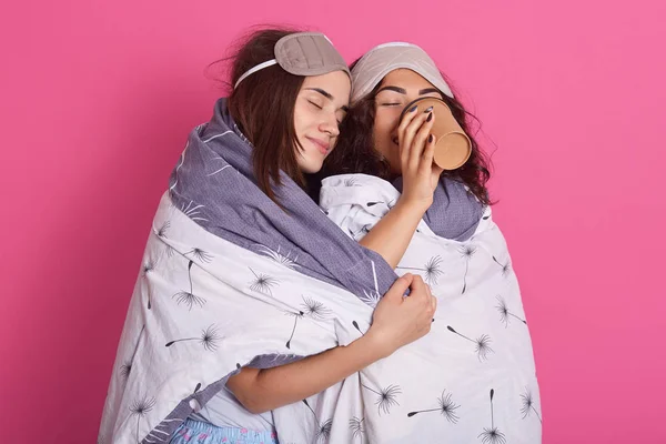 Studio shot of woman drinking coffee while her friend with closed eues hugs her during pajamas party, friends wearing blanket and has blindfolders on heads, posing isolated over pink background. — Stock Photo, Image