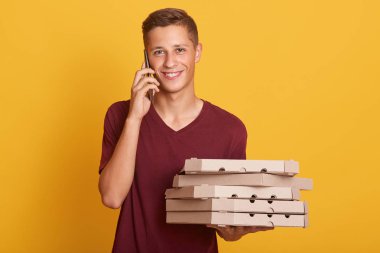 Genç neşeli çocuk evde sarı arka plan üzerinde izole ayakta, elinde pizza ve akıllı telefon ile karton kutular tutarak, konuşma sahip, telefonda konuşurken, doğrudan kamera bakıyor.