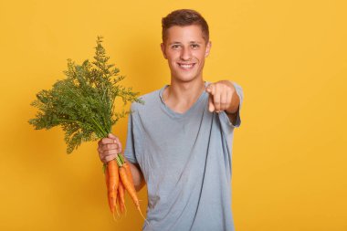 , Bir taraftan olgun havuç demet tutan, kamera atucu ile işaret jest yapma, kol yetiştirmek neşeli hoş genç adam portre, doğrudan kamera bakıyor. Duygular konsepti.