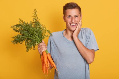 El ile yüzüne dokunmadan yakışıklı faihbroadcasting genç adam yatay çekim, güzel yüz ifadesi, bir yandan havuç demet tutarak, stüdyoda sarı arka plan üzerinde izole poz.