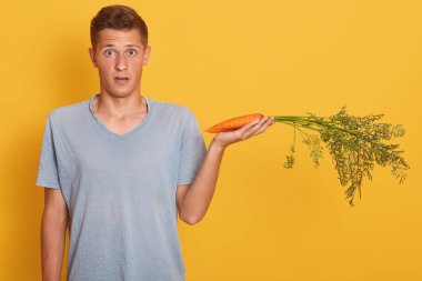 Onun ağzına ve gözleri şok ile açılış etkilendim tatlı genç adam resmi, elinde bir havuç tutan, sürpriz olmak, sebze düşkün, stüdyoda sarı arka plan üzerinde izole poz.