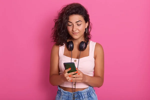 Retrato de linda adorable mujer de pie aislado sobre fondo rosa en el estudio, escribiendo mensajes, siendo adicto a los sitios de redes sociales, tener auriculares alrededor del cuello. Concepto Juventud y Red . — Foto de Stock