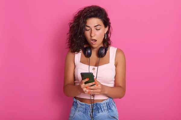 Image of impressed shocked young lady opening her mouth widely with shock, looking at device screen attentively, watching impressive content, wearing pink top and jeans. Technologies concept. — Stock Photo, Image