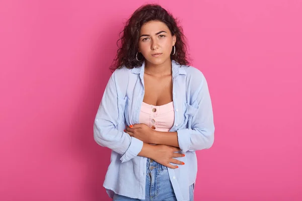 Portrait of suffering tired young woman keeping hands at stomach, having stomachache, feeling uncomfortable, being unhealthy, looking miserable, wearing casual clothes. People and health concept.