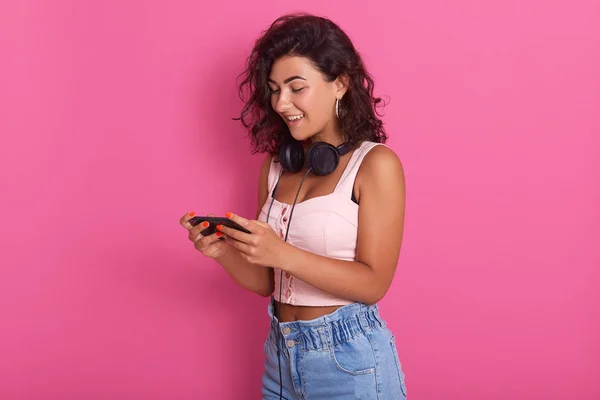 Positief oprecht model dragen van accessoires, lichte top en jeans, druk met haar apparaat, het houden van mobiele telefoon in handen, op zoek ontspannen, surfen sociale netwerksites, het spelen van games, plezier. — Stockfoto