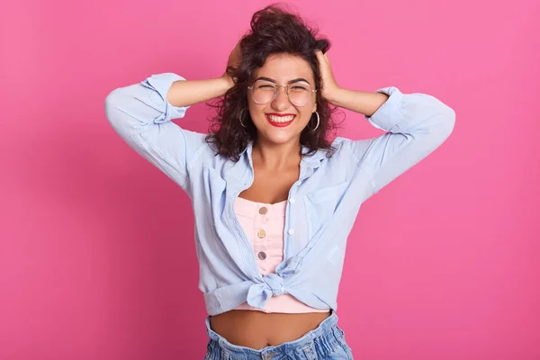 Bild av glad ung vacker kvinna som står isolerad över rosa bakgrund i studion, hålla händerna på huvudet, skrattar uppriktigt, bär jeans, skjorta och rosa topp. Fri tid koncept. — Stockfoto