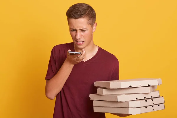 Retrato de mensajero joven insatisfecho teniendo una conversación desagradable con el cliente por teléfono, sosteniendo cajas con pizza, confundiéndose con la ubicación, mirando sceen dispositivo. Concepto de entrega de pizza . — Foto de Stock