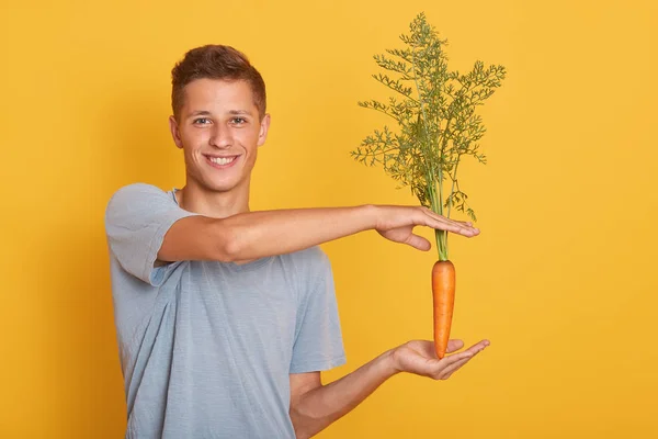 Horizontal shot of slender charming youngster smiling sincerely, making gesture, showing size of carrot, holding raw orange ripe vegetable, stick to healthy diet. People and healthy food concept.