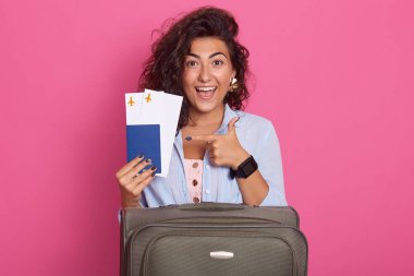 Bavullu bir kadının stüdyo çekimi, elinde uçak biletini ön parmakla gösteren, pembe arka plan üzerinde izole edilmiş, dişli gülümsemeli model, coşkulu mutluluk, wearinf şık kıyafeti.