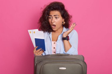 Koyu kıvırcık saçlı, ağızaçık, pasaport tutan, biniş kartı bileti tutan, belgelerine şok edici bir ifadeyle bakan, pembe arka planda izole edilmiş, rahat kıyafetler giyen genç kadın.
