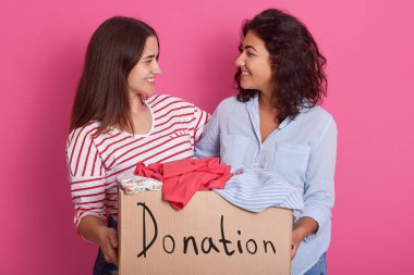 Close up portrait of two girls volunteering, women holding box with clothes for poor people, attractive female making donation, ladies posing and looking at each other. People and charity concept. clipart