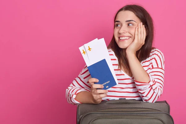 Wizerunek kobiety w paski koszula holdingh paszport i bilety lotnicze, siedzi w pobliżu walizki, marzy o przyszłej podróży, patrząc na bok. Skopiuj miejsce na reklamę lub tekst promocyjny. Koncepcja podróżowania. — Zdjęcie stockowe