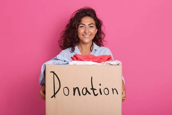 Picture of good looking energetic woman with black curly hair smiling sincerely, doing good things, being fond of charity, having kind heart, following peaceful lifestyle, holding box with donation.