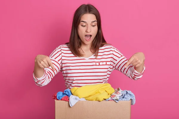 Picture of enthusiastic positive volunteer making gestures, pointing to carton box with forefingers, opening her mouth widely, wearing stripped sweatshirt, looking at various items of clothes. — Stock Photo, Image