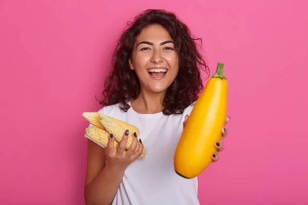 Horizontal shot of cheerful attractive lady looking directly at camera with toothy smile, holding corn cobs and zucchini, having dark curly hair, posing isolated over pink background in studio.