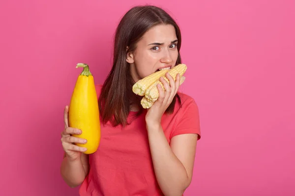 Close up portrait of hungry brunette model posing isolated over pink background in studio, holding bright yellow squash and corn cobs in both hands, eating corn. People and healthy diet concept.