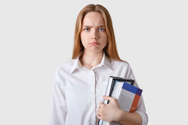 Studio shot of dissatisfied young brown haired woman looking directly at camera, posing over white background, angry girl wearing white blouse, holding paper folder, showing negative attitude. — Stock Photo, Image
