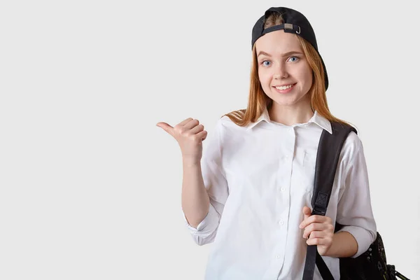 모자, 흰색 블라우스, 배낭을 입은 매력적인 여학생의 초상화를 클로즈업하고 엄지 손가락으로 흰색 배경에 고립 된 것을 가리킵니다. 프로모션 텍스트 또는 광고용 공간 복사. — 스톡 사진