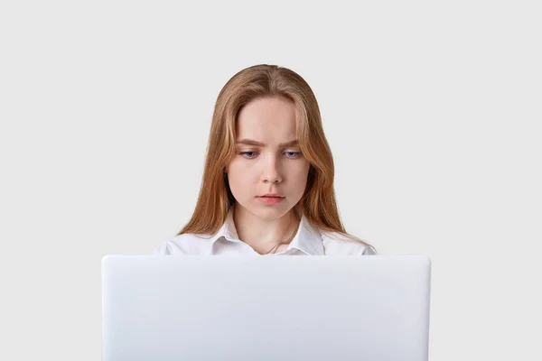 Vista frontal de la niña adulta concentrada sentada frente a un portátil blanco, que tiene trabajo en línea, mira a la pantalla del ordenador, usando blusa, tiene cabello liso y claro, utilizando Internet inalámbrico . —  Fotos de Stock