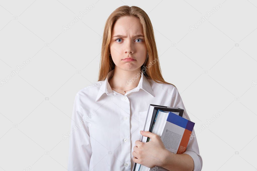Studio shot of dissatisfied young brown haired woman looking directly at camera, posing over white background, angry girl wearing white blouse, holding paper folder, showing negative attitude.