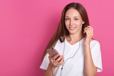 Kulaklıklara dokunan gülümseyen kadının stüdyoyatay çekimi, beyaz rahat tişört giyen, en sevdiği müziğin tadını çıkaran, kameraya gülümseyen beyaz tenli beyaz genç kadın. Kopyalama alanı.