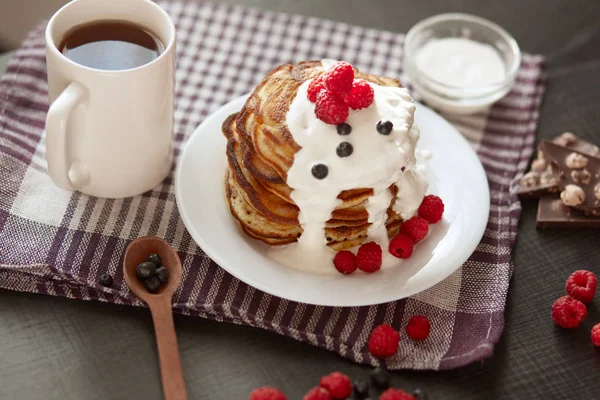 image of home made pancakes with sour cream on white plate decorated with berries, fresh blueberries and raspberries, cup of tea or coffee, spoon, pieces of chocholate on cotton checkered towel.