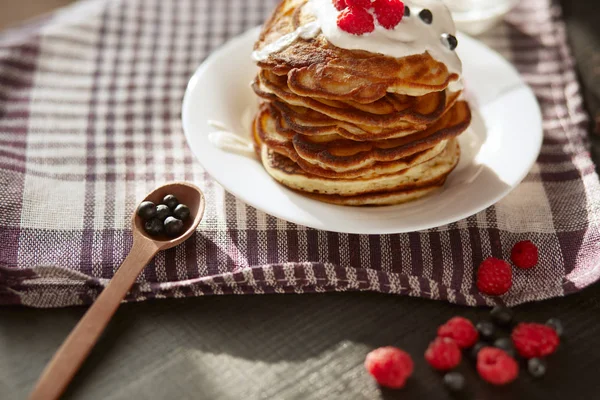 Image of cheese pancakes with sour cream and fresh berries, blackberry jam in wooden spoon, whipped cream on muffins top, served in white plate on with kitchen checkered dish towel, tasty dessert. — Stock Photo, Image