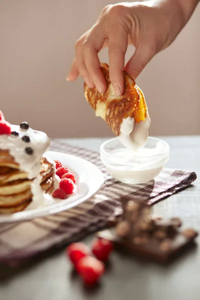 Breakfast set concept. Faceless photo of someone's hand dipping pancake in sour cream. Pancake tower with fresh raspberries and blueberries, sour cream on porcelain plate over dark wooden table.