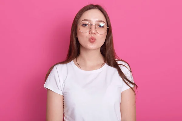 Bedövning kvinnlig modell poserar med Kiss Face uttryck på Rosy Round. Närbild porträtt av snygg europeisk flicka stående framför Studio Wall, klädd casual vit t-shirt och glasögon. — Stockfoto