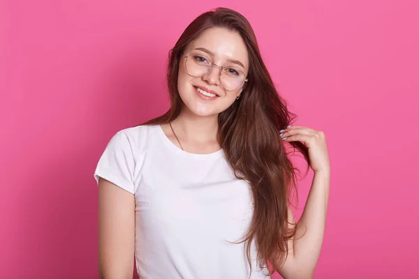 Mulher sorridente em óculos posando em estúdio contra a parede rosa olhando diretamente para a câmera, inclina a cabeça e toca sua meada de ouvir, tem aparência agradável, estar de bom humor, vestido t-shirt branca . — Fotografia de Stock