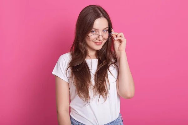 लांब केस असलेल्या आकर्षक तरुण स्त्रीच्या घरातील शॉट चष्माच्या रिमवर हात ठेवते, प्रासंगिक उपकरणे परिधान करते, गुलाबी स्टुडिओच्या भिंतीविरूद्ध मॉडेल थेट हसत दिसते. लोक संकल्पना . — स्टॉक फोटो, इमेज