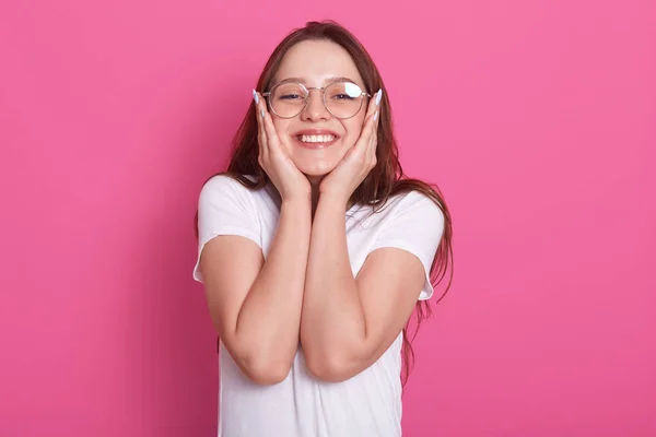 Imagem de uma jovem feliz cheia de esperança e atitude positiva olhando para a câmera com um sorriso alegre e mantém as mãos nas bochechas, posando sobre fundo de estúdio rosa, vestida com roupas casuais e óculos . — Fotografia de Stock