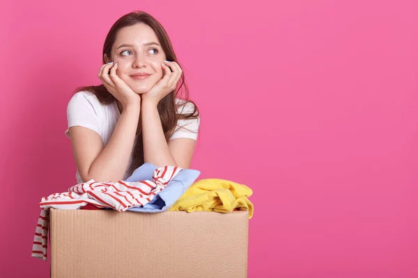 Horizontal shot of cheerful young volunteer standing isolated over rosy studio background, posing near carton box full of donated clothes. Smiling woman looking aside, keepa hands under chin. — Stock Photo, Image