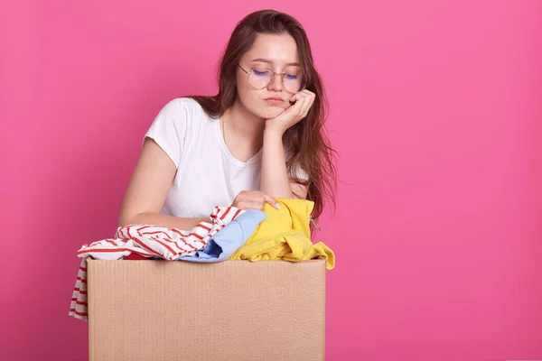 Close up portrait of unhappy young woman posing with box of clothes donation, has sad facial expression, looking down, keeps hand under chin, packing clolothes for secondary use for poor people. — Stock Photo, Image